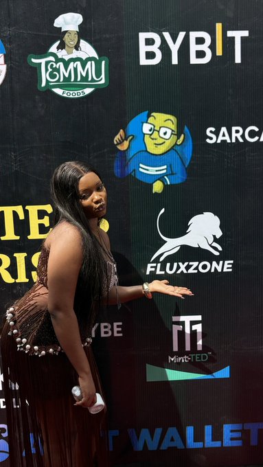A woman in a brown fringed dress poses in front of a backdrop with various sponsor logos including 'TOMMY FOODS', 'BYBIT', 'FLUXZONE', and 'MintTED'.