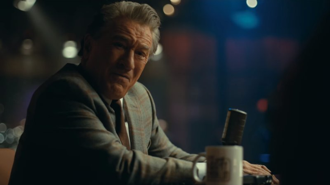 An older man in a plaid blazer sits at a dimly lit bar with a mug in front of him, looking toward someone off-camera.