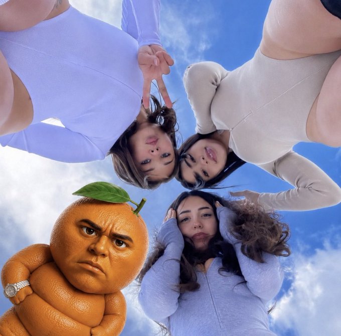 Four young women lean in from above making peace signs against a cloudy blue sky, while an Annoying Orange character with a grumpy human-like face poses in the lower left corner. The image ties into the post's meme-style promotion of the "$Udin" token, using the Annoying Orange's "annoying" persona as playful branding for the crypto project.