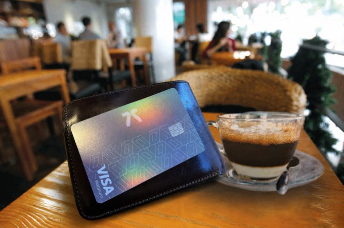 The image features a wallet with a Visa card inside of it placed on top of a table. The wallet has a blue Visa logo, indicating that it's a Visa card holder. There are also two cups on the table - one containing coffee and another filled with ice cream. A chair is visible in the background, suggesting that this scene takes place in an indoor setting like a cafe or restaurant.