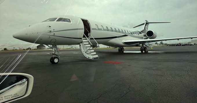 A white private jet parked on a tarmac with its airstair deployed, photographed from inside a vehicle with the side mirror visible in the lower left corner.