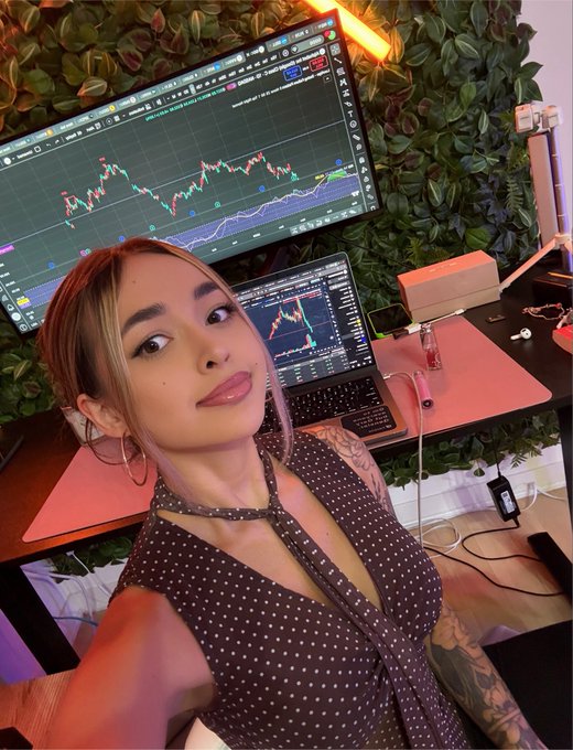 A woman poses for a selfie at her trading setup, with multiple monitors displaying candlestick charts behind her. The post's caption references "$FEFER" and "@Stable," suggesting she's a trader waiting on resolution of a position involving that token.
