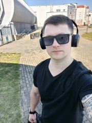 A selfie of a young man wearing black sunglasses and large black headphones, standing outdoors near what appears to be a stadium.