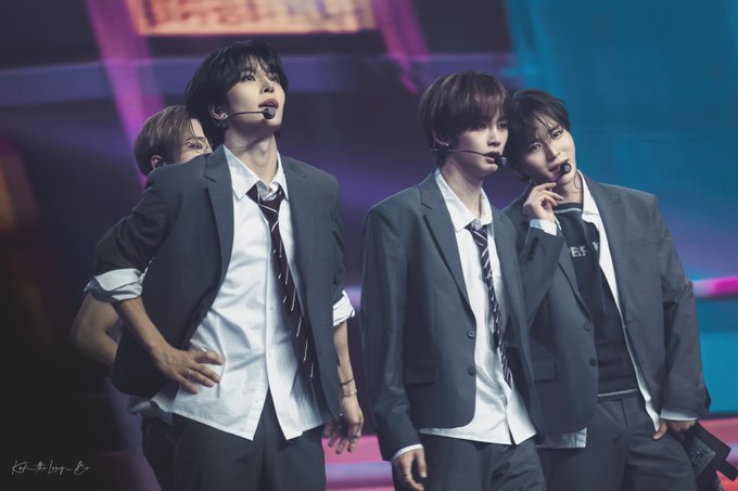 Four performers in dark suits, white shirts, and loosened striped ties pose on a concert stage wearing headset microphones under blue and pink lighting, matching the "#ALPHADRIVEONE #ALD1" tour and showcasing member ZHOU ANXIN (周安信).