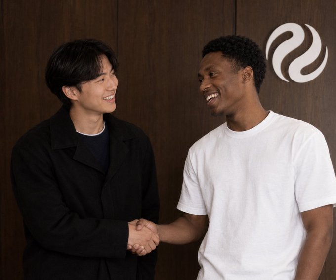 Two smiling young men, one East Asian wearing a black jacket and the other Black wearing a white t-shirt, shake hands in front of a wooden wall featuring a stylized white logo.