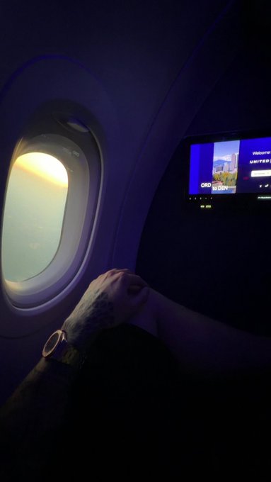 A passenger's tattooed hand rests near an airplane window at sunset, with a seatback screen displaying a 'UNITED' flight information page in purple cabin lighting.