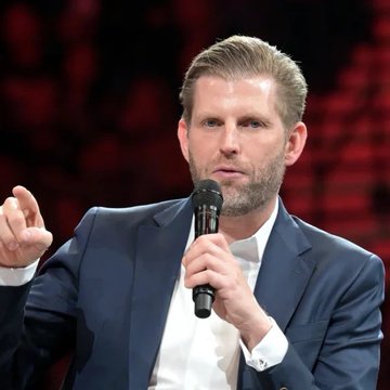 Eric Trump, holding a microphone and gesturing while speaking at an event, discusses his claim that the SWIFT financial system is broken and will be replaced by cryptocurrency.