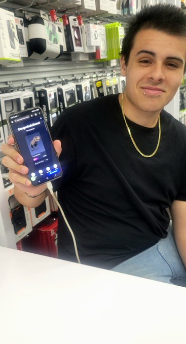 A young man wearing a black t-shirt and gold chain sitting in what appears to be a phone accessories store, holding up an iPhone showing a social media profile while smiling at the camera.