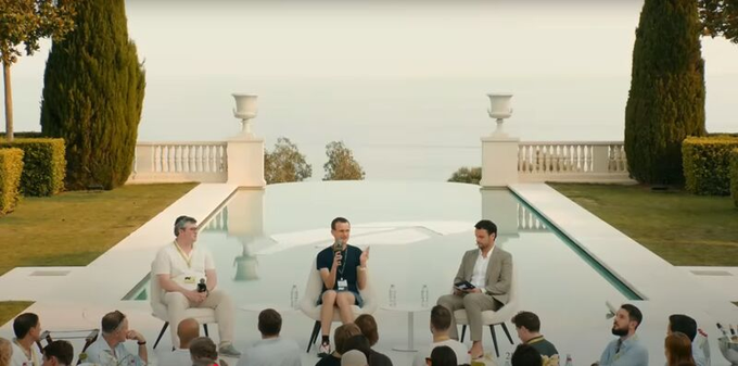 Three men sit on stools on an outdoor stage by a pool overlooking the sea, appearing to participate in a panel discussion at what looks like a luxurious seaside venue, likely related to the Arbitrum, Robinhood, and Ethereum collaboration mentioned in the post.