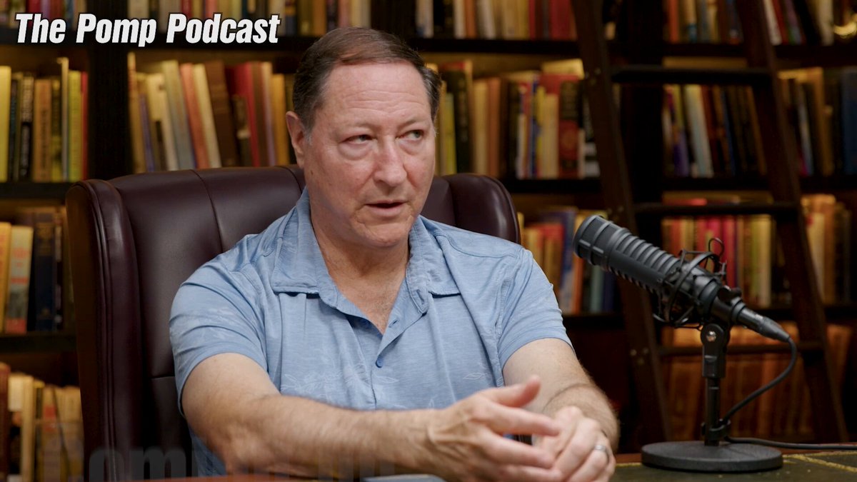 The image features a man sitting at a desk with a microphone, likely recording an interview or podcast. The background shows numerous bookshelves filled with various books, suggesting that this might be a library or a room dedicated to reading and learning. The man is wearing a blue shirt and appears to be in the process of speaking into the microphone.