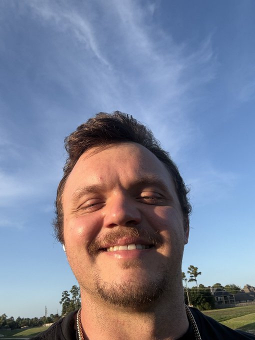 A close-up outdoor selfie of a smiling man with a mustache wearing a black shirt and a silver chain, taken against a bright blue sky with scattered clouds.