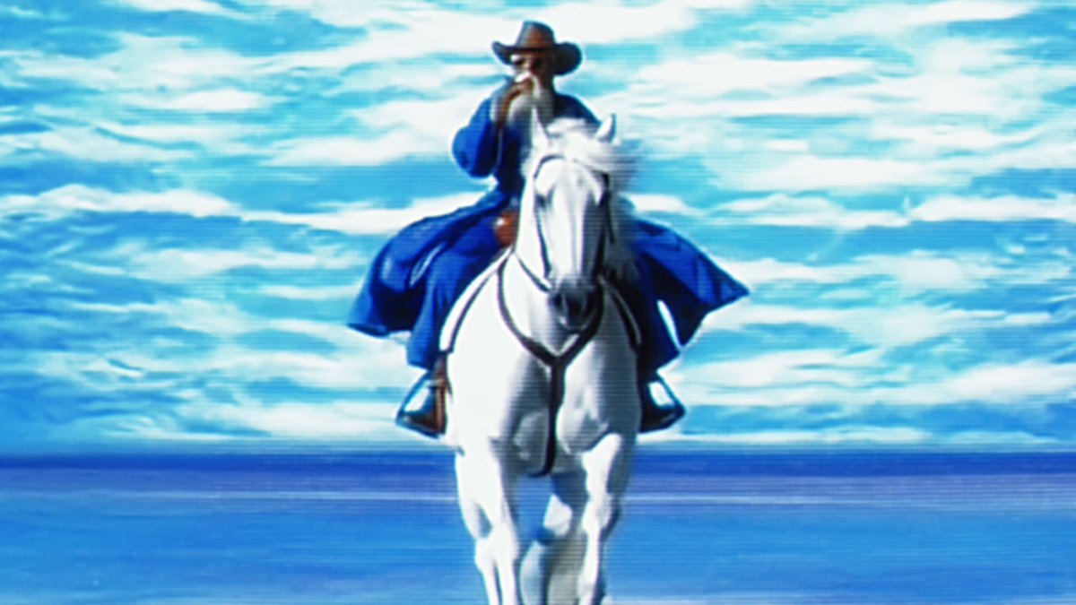 The image features a man riding a white horse, with his back turned towards the camera. He is wearing a blue coat and hat, which contrasts with the white horse. The background of the image shows an ocean or sea, adding to the overall ambiance of the scene. There are no other objects in the image besides the man on the horse and the water.