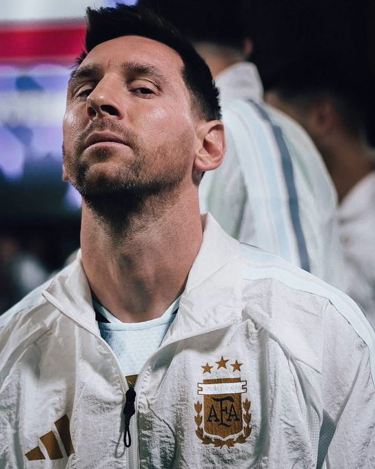 A close-up portrait of Argentine soccer player Lionel Messi wearing a white Argentina national team jacket featuring the AFA crest with three stars above it.