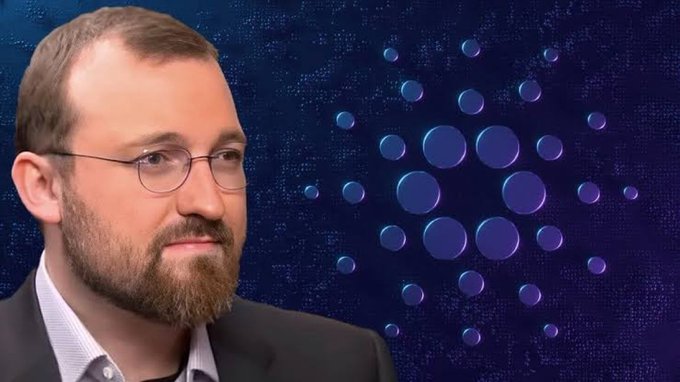 A portrait of a bearded man with round glasses wearing a suit, set against a dark blue background featuring a circular pattern of glowing blue dots resembling the Cardano cryptocurrency logo.