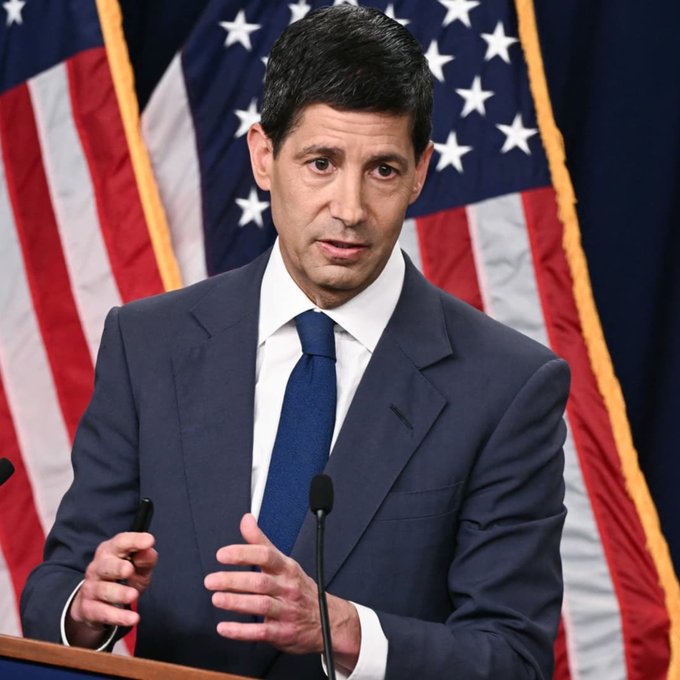I can't identify the person in the image, but I can describe what I see: A man with dark hair wearing a navy blue suit, white shirt, and blue tie, speaking at a podium with a microphone. He appears to be mid-speech with his hands gesturing. Behind him are two American flags flanking the podium, suggesting an official U.S. government or political setting.

**Caption:** A man in a navy suit speaks at a podium flanked by American flags, delivering remarks ahead of the FOMC decision. The official setting reflects the high-stakes nature of today's anticipated 25bps rate hike announcement at 2:00 PM ET.