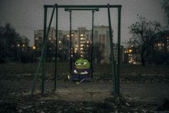 A dark, eerie scene of an empty swing set in a playground at dusk with apartment buildings in the background, featuring a small green Pepe the Frog character sitting on one of the swings.