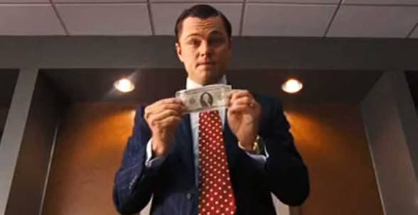 Leonardo DiCaprio as Jordan Belfort in 'The Wolf of Wall Street' holds up a single dollar bill while wearing a blue suit and red polka-dot tie.