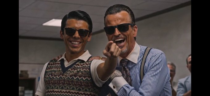 Two smiling men in vintage attire wearing sunglasses pose together in an office setting, with one pointing toward the camera.