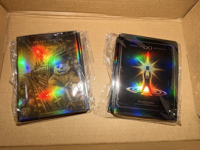 A box containing sealed holographic packs of "$FUZZY ON XRP" trading cards, featuring artwork of astronauts on the packaging.
