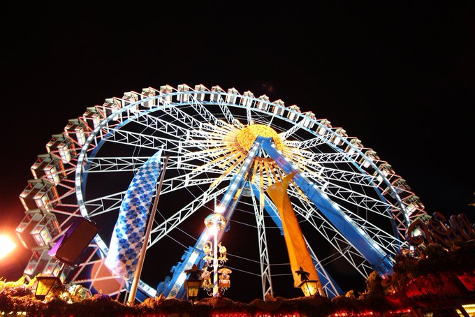 The image features a large Ferris wheel at night with blue and white lights illuminating its structure. The Ferris wheel has multiple levels and appears to be an attraction at a fair or amusement park, as indicated by the presence of flags on top. The photo is taken from below, looking up towards the Ferris wheel, which emphasizes its height and grandeur against the dark sky.