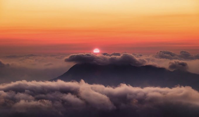 A breathtaking sunrise (or sunset) with a vibrant orange and red sky, where the sun appears as a small bright orb emerging above a sea of clouds and the silhouette of a mountain peak.