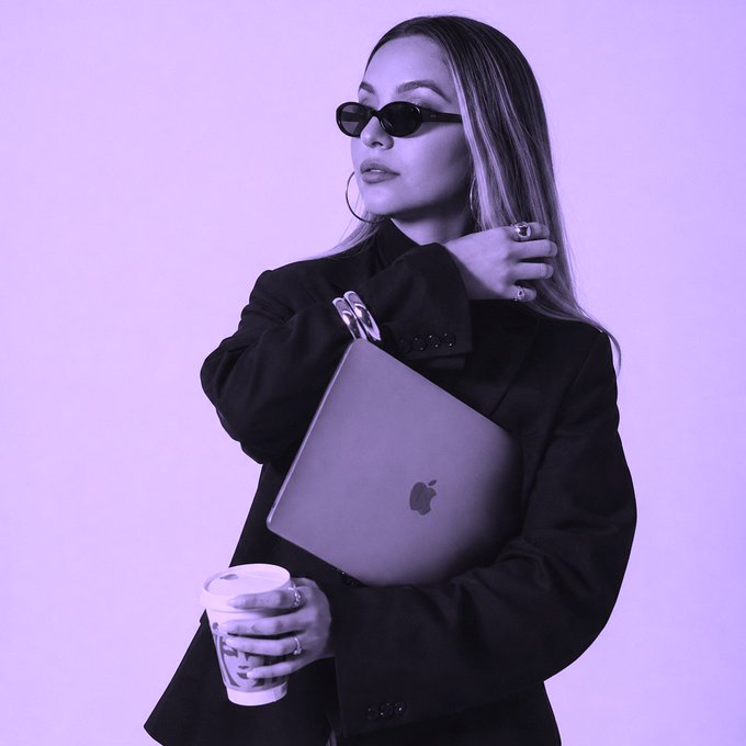 Woman in a black blazer and sunglasses holding a silver Apple laptop and a coffee cup, photographed with a purple color filter.