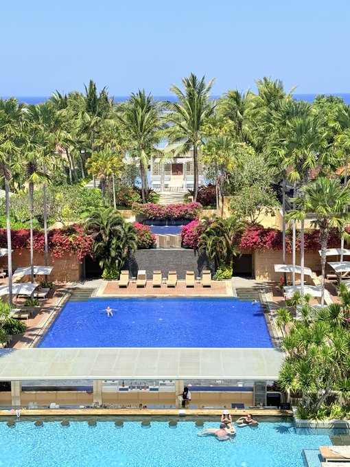 A luxurious tropical resort features two stunning pools surrounded by lush palm trees and vibrant pink bougainvillea, with the ocean visible in the background, capturing the serene and sun-drenched paradise vibe of a Bali getaway.