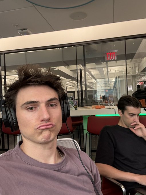 Two young men sit in an office space wearing headphones, with the foreground subject giving a pouty expression toward the camera while the man behind him rests his chin on his hand, both appearing to prepare for a live stream. An illuminated red "EXIT" sign is visible through the glass partition behind them, fitting the casual workspace vibe of their upcoming Gemini stream with special guest Alex Needleman, CEO of Dyli.