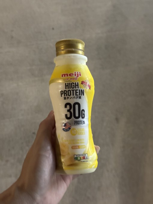 A hand holding a Meiji CP-Meiji High Protein banana-flavored bottle advertising 30g of protein with no added sugar.