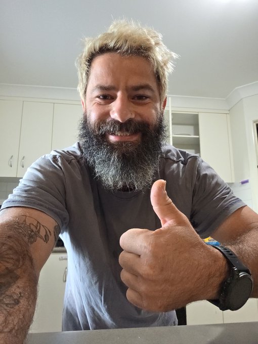 A man with bleached blonde hair and a thick, graying beard smiles and gives a thumbs-up while taking a selfie in a kitchen, wearing a gray t-shirt and a black smartwatch.