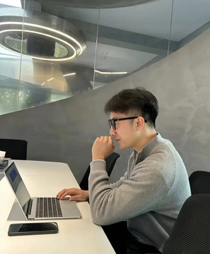 A young man wearing glasses and a gray sweater sits at a desk using a MacBook laptop in a modern office setting with a circular ceiling light.