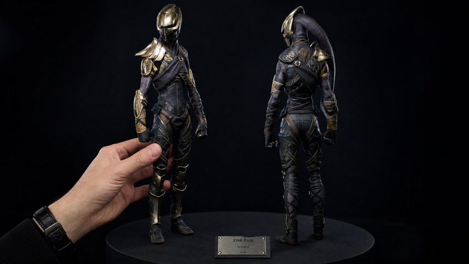 A detailed action figure of a character in ornate gold and dark armor with a horned helmet, shown from front and back views on a display base, with a hand for scale.