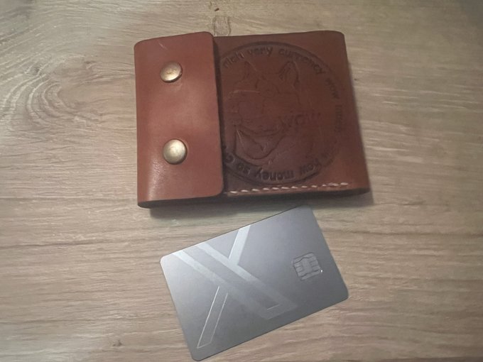 A brown leather bifold wallet with embossed artwork and metal snap closures resting on a wooden surface, positioned above a silver metal card featuring a geometric 'K' or arrow logo.