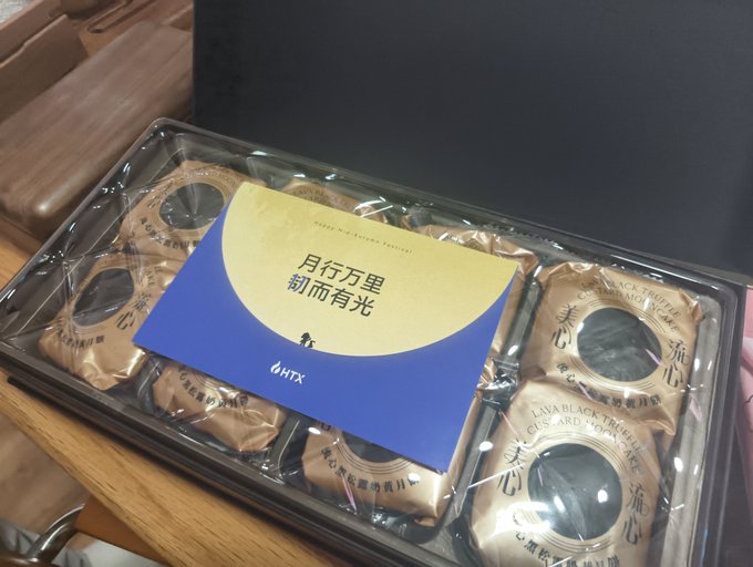 A black gift box containing individually wrapped mooncakes with gold packaging, featuring a card with Chinese text '月行万里 前而有光' and the 'HTX' logo.