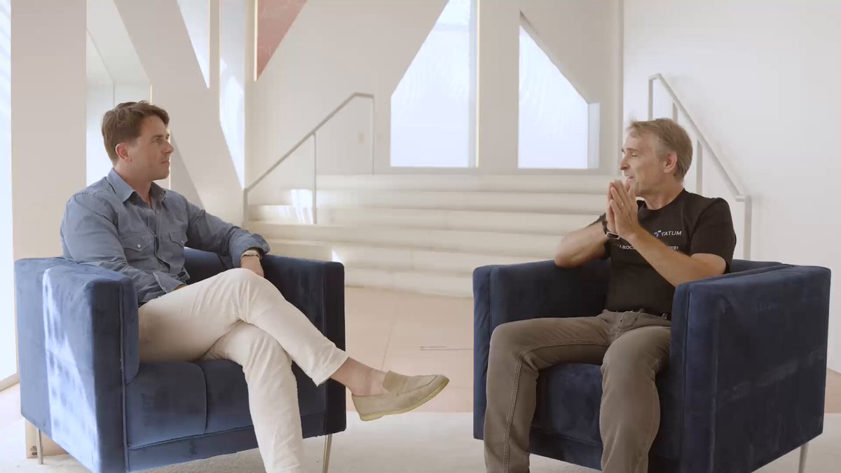 The image shows two men sitting in blue chairs facing each other with their hands clasped together. The room has white walls and a staircase visible behind them. There are no texts or other objects present, so the focus of the image is on the two men engaged in conversation.