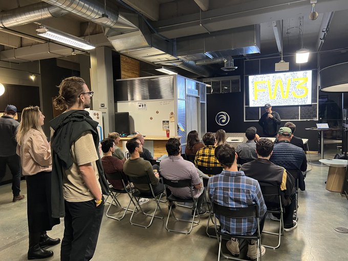 Image 6: A photograph of an indoor tech meetup or presentation in an industrial-style room, with an audience seated and standing while watching a speaker present a slide titled 'FW3' on a large screen.