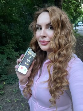 A woman with long wavy reddish-brown hair wearing a pink hoodie holds a phone showing an incoming call screen outdoors.