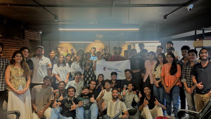 A large group of people poses together indoors, holding a 'Cake Wallet' banner, with 'Cake Wallet' also visible on the wall behind them.