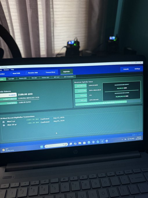 A laptop screen displaying a DigiDollar blockchain wallet interface showing a balance of 2188.00 $DD with transaction history options for Send/Receive DGB, alongside hardware wallets visible on a desk in the background.