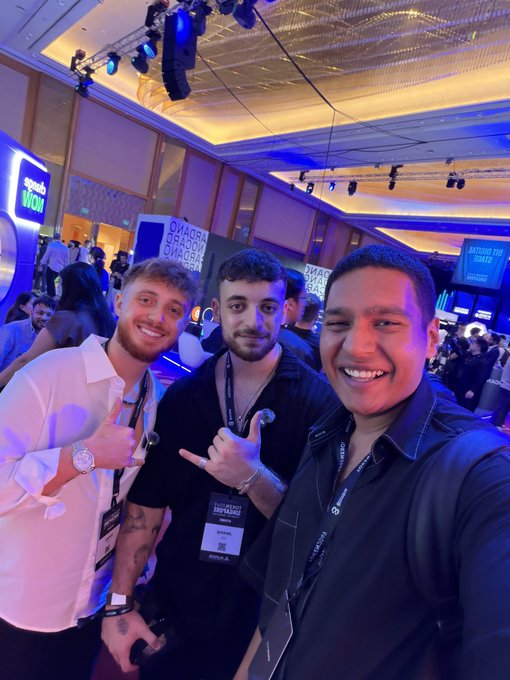 Three young men smile and pose for a selfie at an event with blue lighting, wearing lanyards and event badges in a busy convention hall.