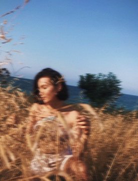 A film-style photo shows a woman with dark hair standing among tall golden grass with the ocean and trees in the background, giving an artistic, dreamy aesthetic.