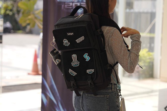 A person wears a black backpack decorated with various patches, including "STOPLOSS" and "TRADE4FUN" stickers, referencing two of the finalists in the UEX Global Alpha Tournament by Bitget. The image cleverly visualizes team support by having the "Stoploss" patch prominently displayed on the backpack.