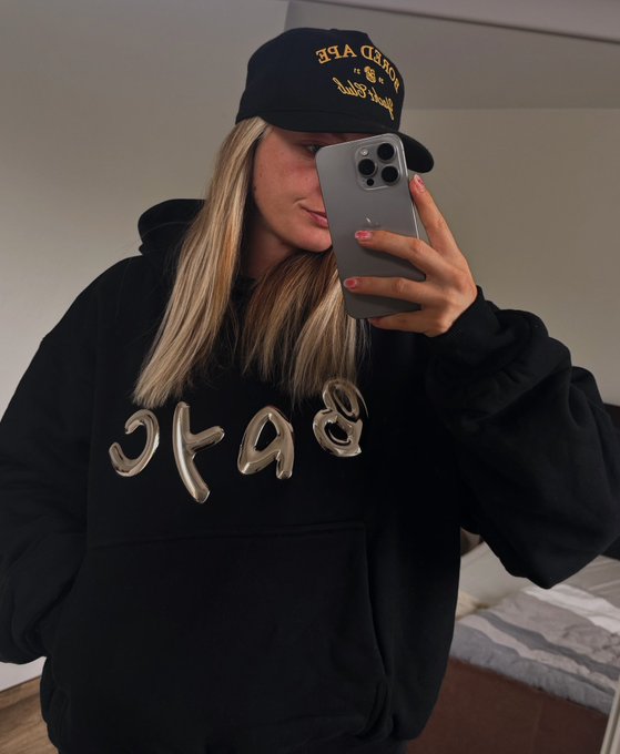 A blonde woman takes a mirror selfie wearing a black 'BAKE' hoodie with graffiti-style metallic lettering and a black 'ROLLER' baseball cap.