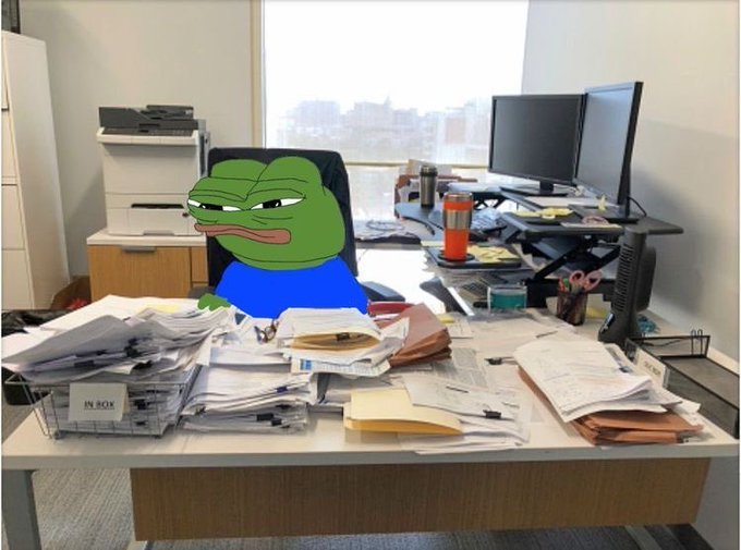 A cartoon Pepe the Frog character (angry expression, blue shirt) sitting at a cluttered office desk piled with paperwork, next to an inbox tray, dual monitors, and a printer.