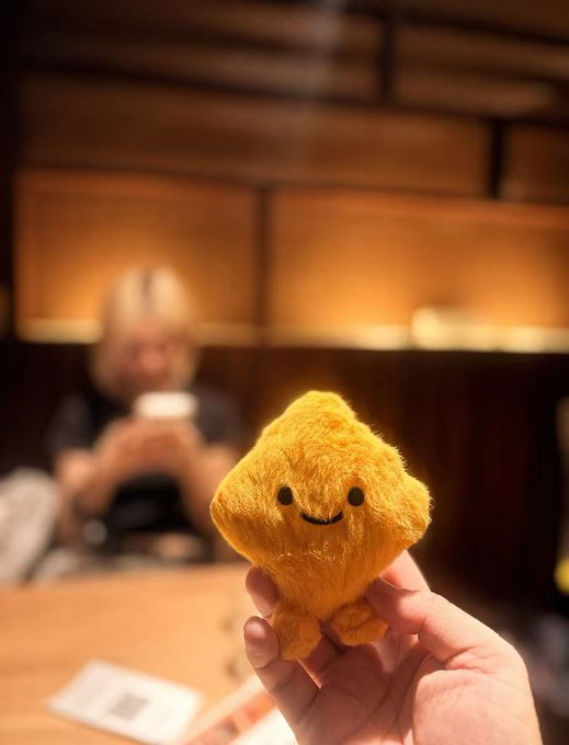 A hand holds up a small, fuzzy, yellow, diamond-shaped plush toy with a smiling face, with a blurred person visible in the background.