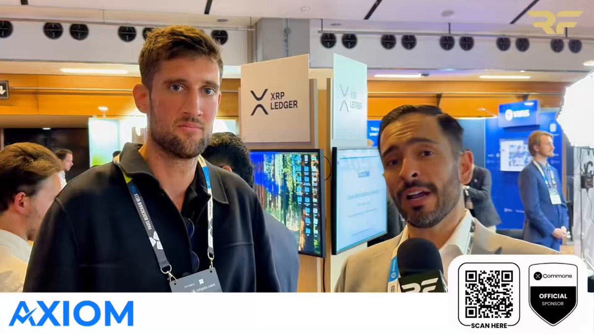 The image features a man wearing a lanyard, standing next to another person who also has a lanyard around their neck. They are both looking at a large screen TV that displays information about an event called "XRP Ledger". The background of the image shows several people in various positions and poses, indicating that this is likely taking place at an event or gathering where attendees can interact with each other and view presentations on the large screen TV.