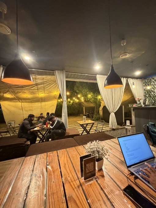 A dimly lit outdoor café or restaurant at night with hanging pendant lights, string lights in the trees, and patrons seated at wooden tables.