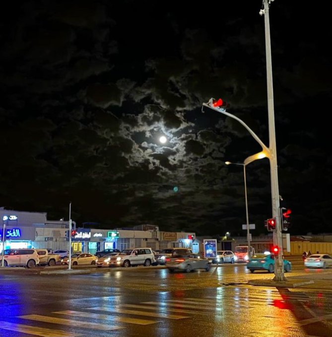 A nighttime city intersection illuminated by traffic lights and storefronts, with the moon visible through scattered clouds in the dark sky.