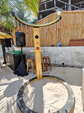 A vintage-style yellow and black photo booth stands on a sandy beach near a bamboo fence and palm trees, with two wooden stools positioned in front of it.