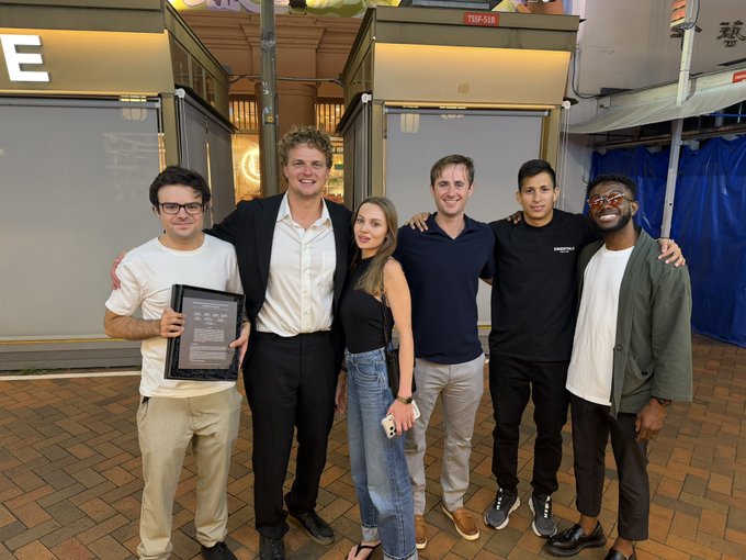 A team of six people pose together at what appears to be a Singapore conference venue, with one member holding a framed award or document. The setting aligns with the Quantum & Privacy Day event mentioned, featuring representatives from NEAR Protocol, Zcash, and Quantus Network advocating against AI surveillance and for private money.